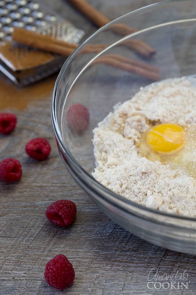 Raspberries and a clear bowl resting on table. The clear bowl is filled with a flour mixture and a freshly cracked egg yolk. 