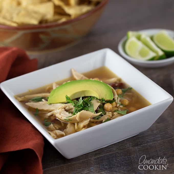 A white square bowl filled with chipotle chicken soup and topped with one slice of avocado.