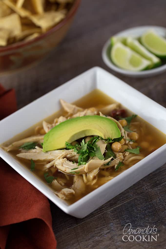 A close up of a square white bowl filled with chipotle chicken soup, topped with one slice of avocado.