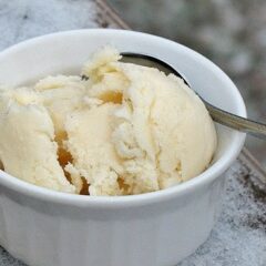 A close up photo of a scoop of homemade vanilla ice cream in a white bowl served with a spoon.