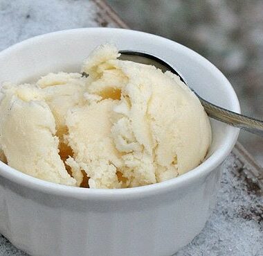 A close up photo of a scoop of homemade vanilla ice cream in a white bowl served with a spoon.