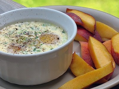 A close up photo of eggs baked in ramekins with herbs on a plate with sliced peaches.