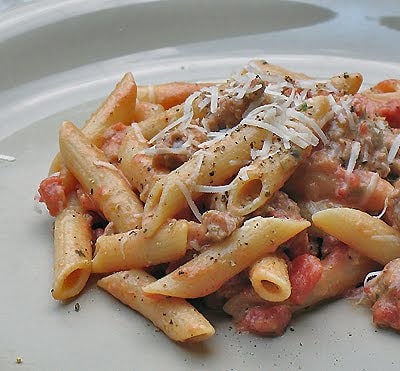 A close up of Penne with tomato sage sausage sauce on a white plate topped with shredded Parmesan cheese.