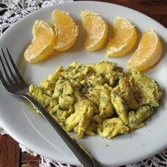 A close up photo of scrambled eggs with garlic, basil and feta with mandarin orange slices to to side on a white plate.