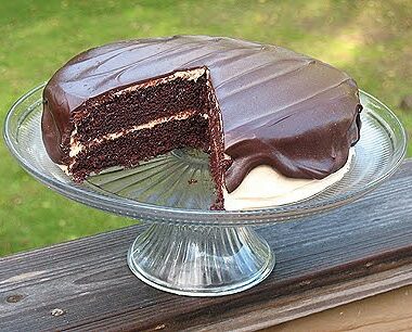 A photo of a chocolate cake with peanut butter frosting and chocolate peanut butter topping resting on a cake stand with a slice removed.