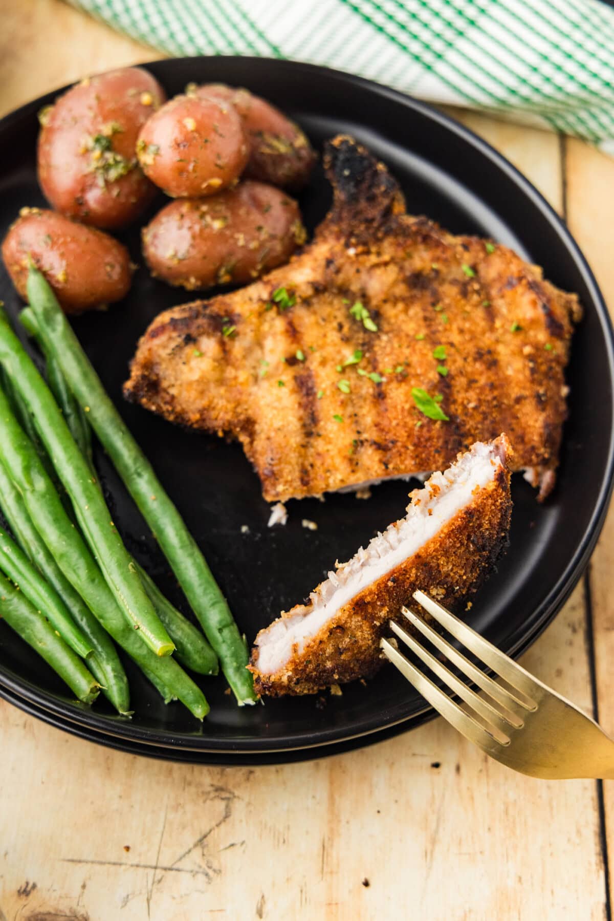 Grilled Breaded Pork Chop on plate with green beans and potatoes and fork lifting a slice