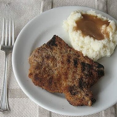 An overhead photo of a grilled breaded pork chop on a white plate with mashed potatoes and gravy and a fork.