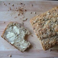 A photo of a loaf of sunflower whole wheat quick bread with a buttered slice to the side all resting on a wooden cutting board.