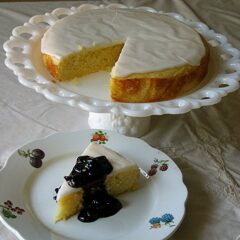 A photo of a lemon cornmeal cake missing a slice on a white cake stand next to a slice of cake on a white plate topped with blueberry sauce.