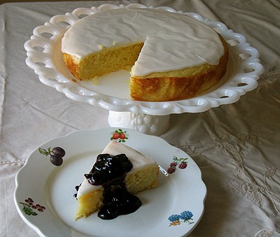 A photo of a lemon cornmeal cake missing a slice on a white cake stand next to a slice of cake on a white plate topped with blueberry sauce.