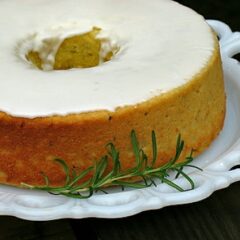 A close up photo of a lemon rosemary olive oil cake resting on a decorative white plate with a rosemary sprig on the side.