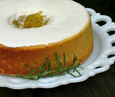 A close up photo of a lemon rosemary olive oil cake resting on a decorative white plate with a rosemary sprig on the side.