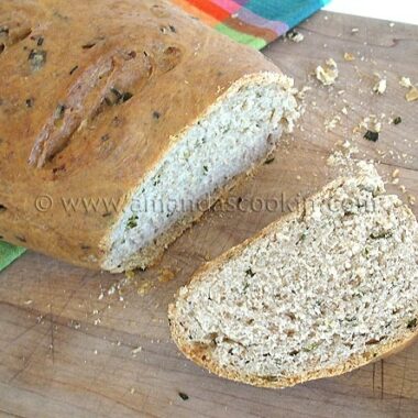 A close up photo of a sliced loaf of scallion chive bread with garlic and rosemary on a wooden cutting board.