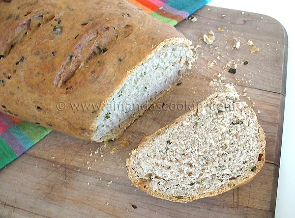 A close up photo of a sliced loaf of scallion chive bread with garlic and rosemary on a wooden cutting board.