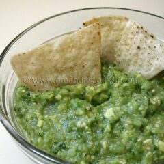 A close up photo of tomatillo onion avocado salsa in a clear bowl with two tortilla chips.