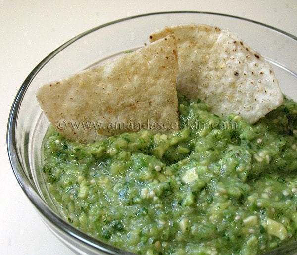 A close up photo of tomatillo onion avocado salsa in a clear bowl with two tortilla chips.