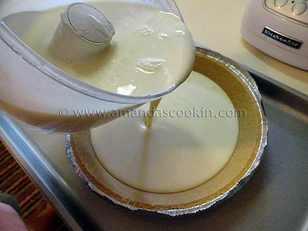 A photo of the ingredients for lemonade cheesecake in a food processor being poured into a graham cracker pie crust.
