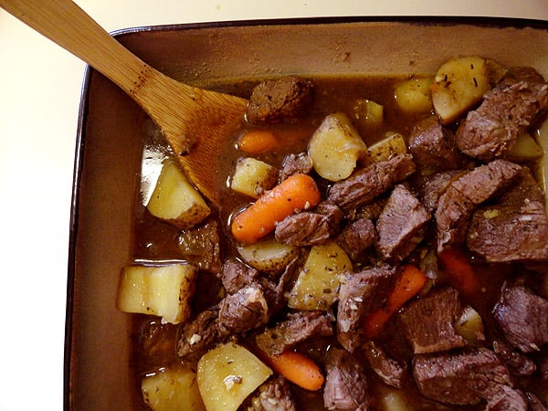 A close up overhead photo of beef stew with parsley, sage, rosemary and thyme in a bowl with a wooden spoon.