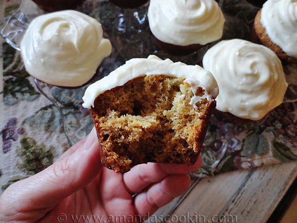 A photo of a half eaten banana cupcake with vanilla cream cheese frosting with whole cupcakes in the background.