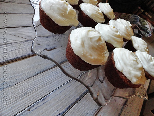 A photo of banana cupcakes with vanilla cream cheese frosting resting on a clear cake stand.
