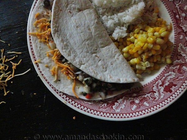 An overhead photo of a steak soft taco resting on a plate with corn and rice to the side.