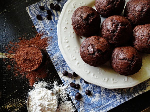 An overhead photo of Starbuck\'s hot cocoa chocolate chip muffins on a white plate with cocoa and flour to the side.