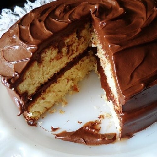 A photo of a classic yellow cake covered in chocolate frosting with a slice removed.