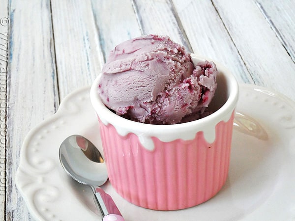 A close up photo of a scoop of vanilla blackberry jam ice cream in a pink bowl with a spoon resting to the side.