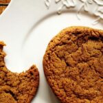 A close up photo of whole sugar topped molasses spice cookie next to a half eaten cookie on a white plate.