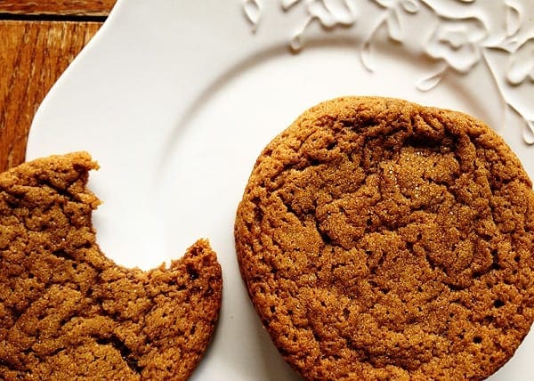 A close up photo of a whole sugar topped molasses spice cookie next to a half eaten cookie on a white plate.