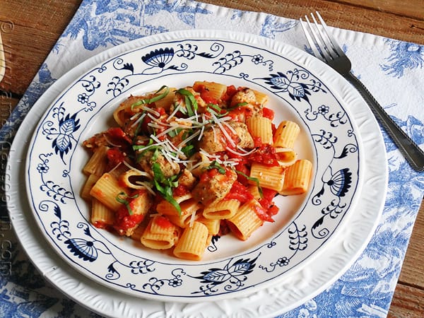 A close up photo of a plate of spiced chicken and mezzi rigatoni topped with grated Parmigiano-Reggiano cheese and fresh basil.