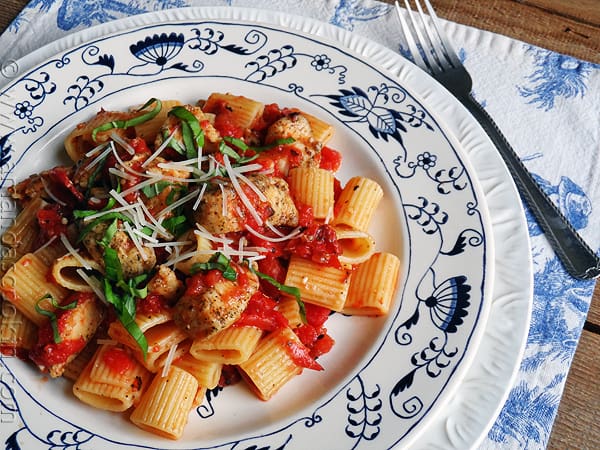 An overhead of a plate of spiced chicken and mezzi rigatoni topped with grated Parmigiano-Reggiano cheese and fresh basil.