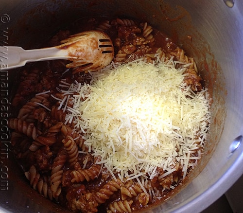 An overhead photo of parmesan cheese over the pasta, sauce and sausage mixture in a bowl.