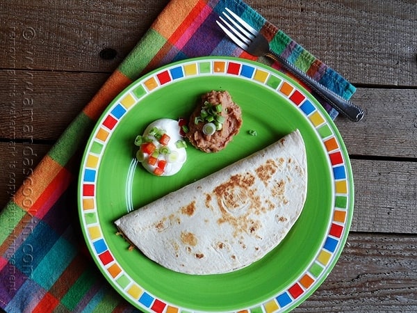 An overhead photo of a beef and bean quesadilla on a green plate with sour cream and refried beans on the side.