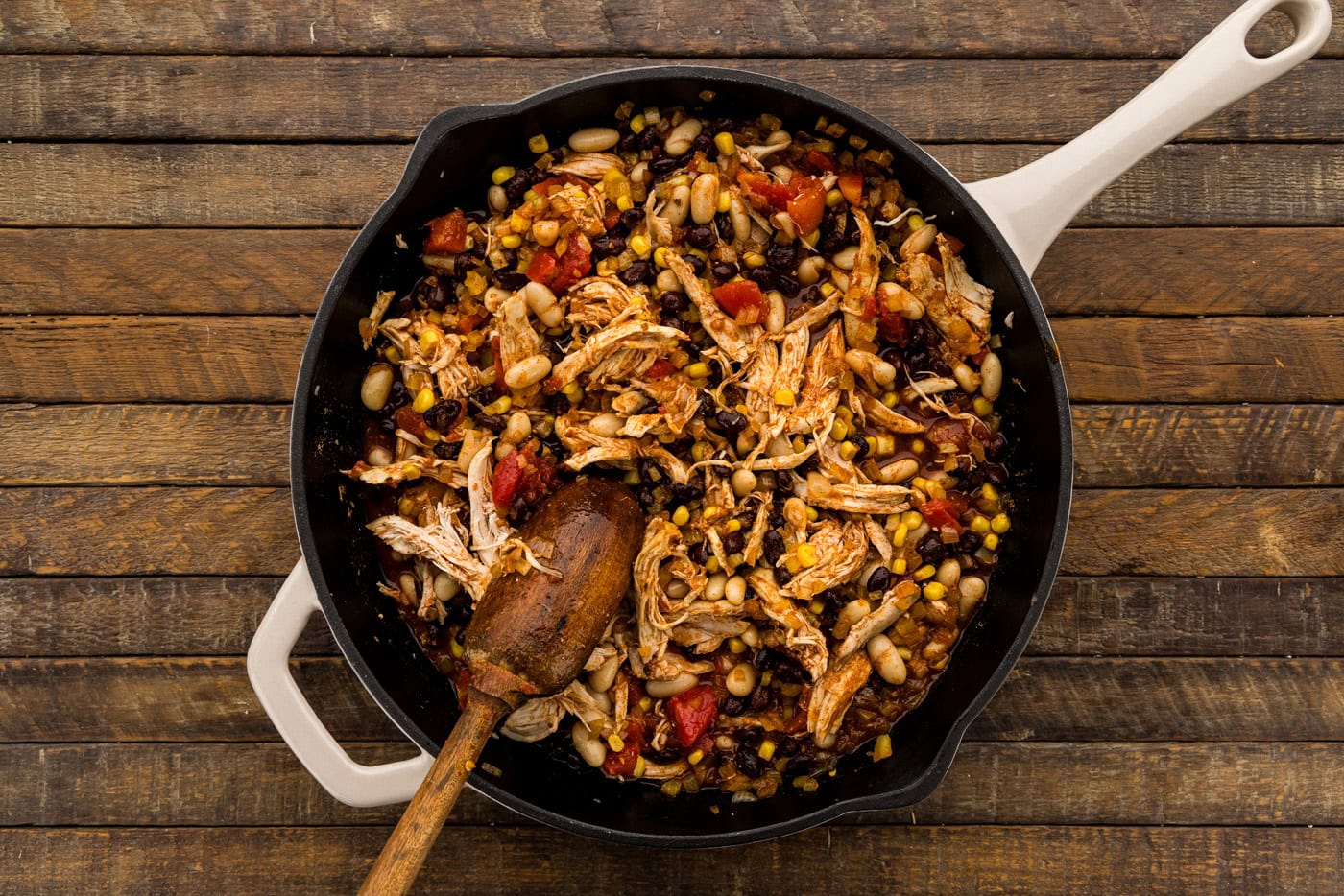 Wooden spoon stirring shredded chicken, beans and corn together in the skillet