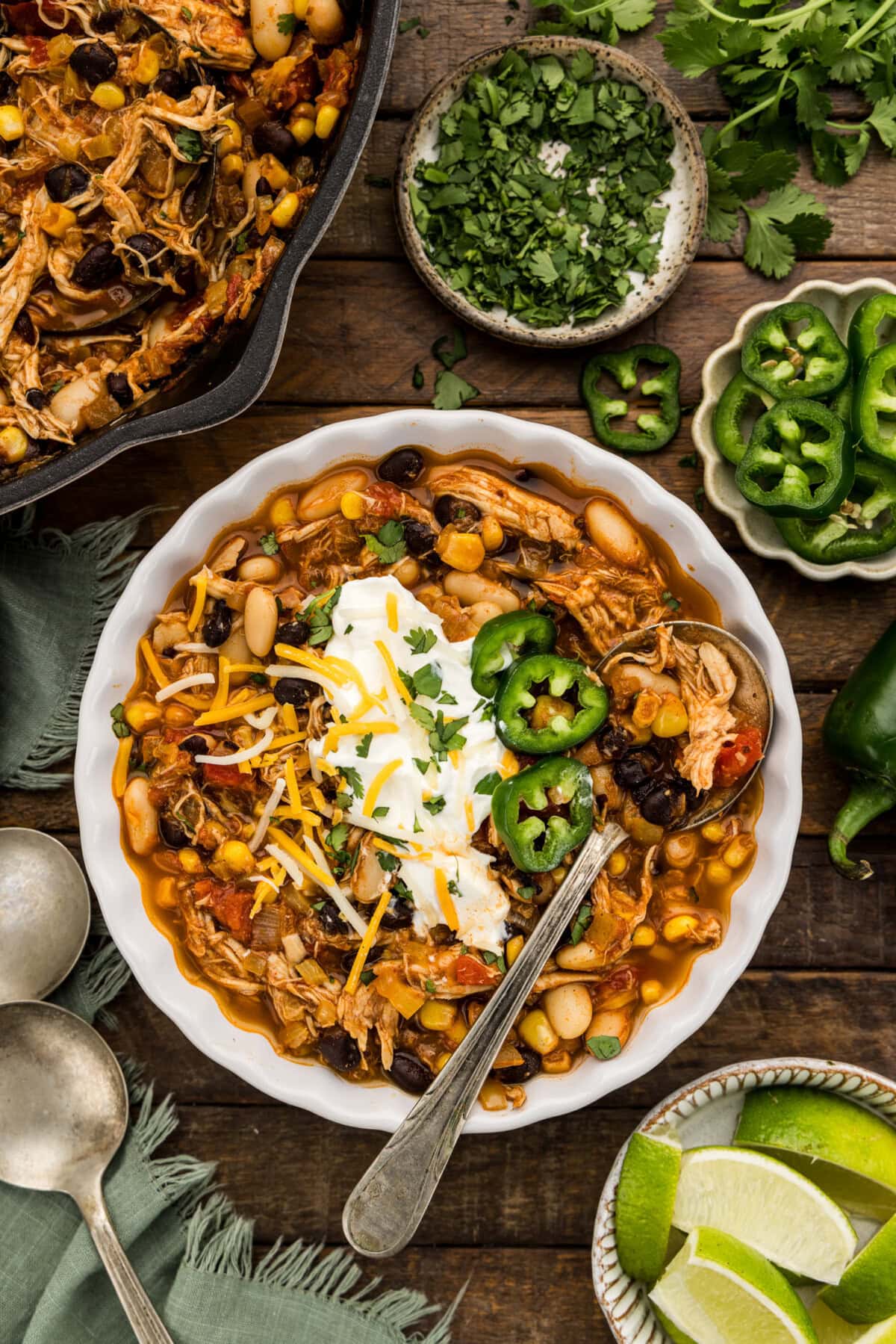 Bowl of black bean chicken chili topped with sour cream, shredded cheese, jalapeño slices and cilantro with lime wedges on the side