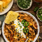 Bowl of chicken chili with black beans and corn topped with sour cream, jalapeños and cheddar with a wedge of cornbread