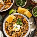 Two bowls of chicken chili with black beans and corn topped with sour cream and jalapeños, served with cornbread, lime wedges, cilantro and iced tea