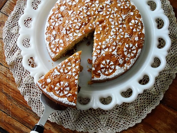 A overhead photo of a Merryfield apple cake on a decorative white platter with a slice being taken out. 