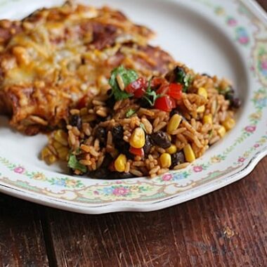 A close up photo of Spanish rice with black beans and corn on a plate with enchiladas in the background.