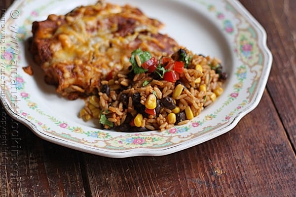 A close up photo of Spanish rice with black beans and corn on a plate with enchiladas in the background.