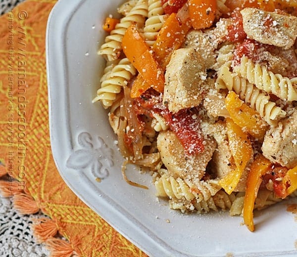 A close up overhead photo of chicken with peppers and pasta in a white bowl topped with Parmesan cheese.
