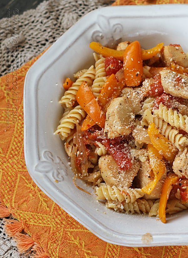 A close up overhead photo of chicken with peppers and pasta in a white bowl topped with Parmesan cheese.