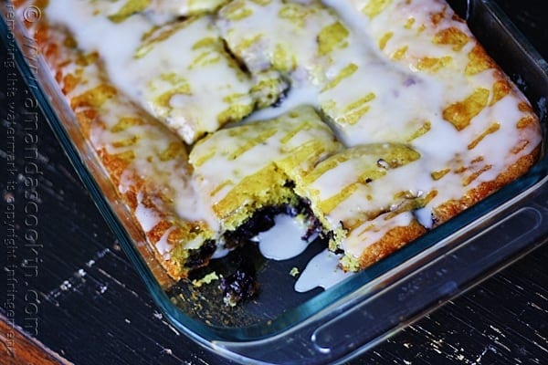 A close up photo of glazed lemon blueberry cake in a dish with a piece removed.