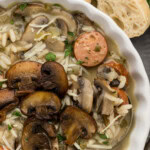 Close up of a bowl of sausage wild rice soup topped with browned mushrooms, grated Parmesan and parsley beside crusty bread