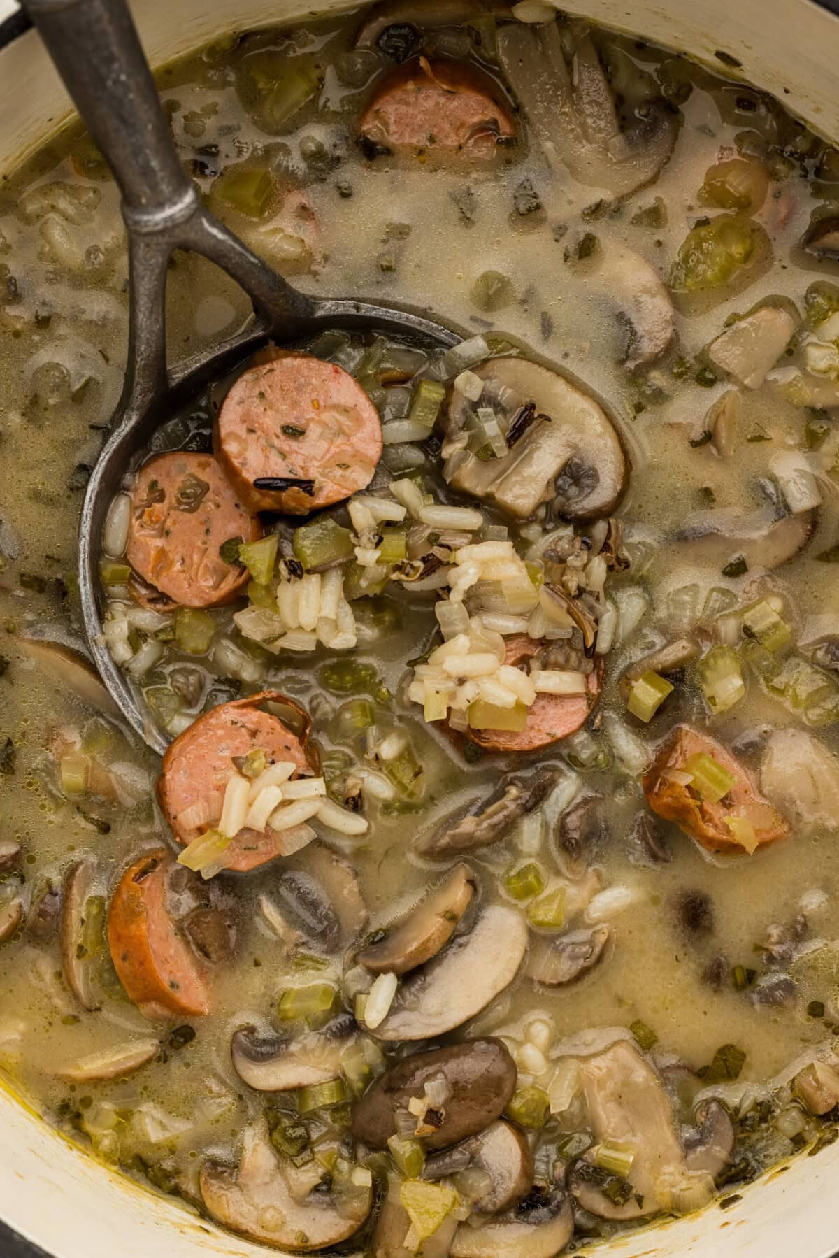 A ladle lifting sliced sausage, mushrooms and wild rice out of a pot of soup