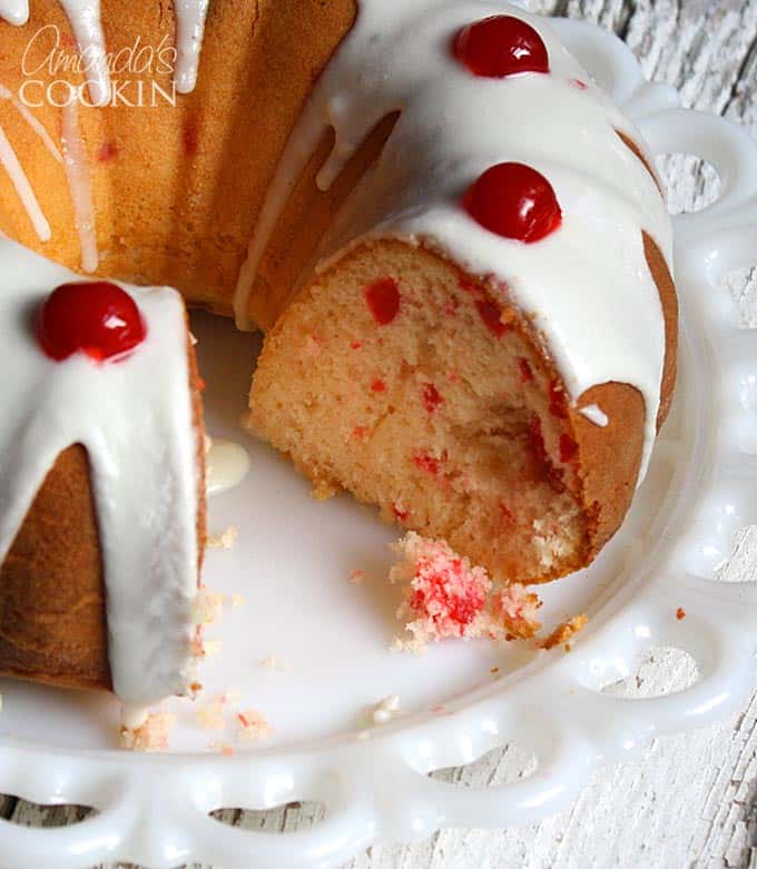 A close up photo of a maraschino cherry Bundt cake on a decorative platter with almond butter glaze and  cherries on top with a piece missing.