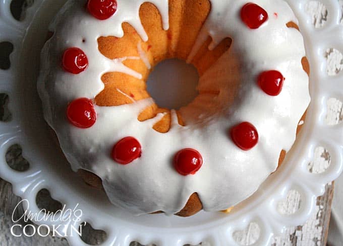 A overhead photo of a maraschino cherry Bundt cake on a decorative platter with almond butter glaze and  cherries on top.