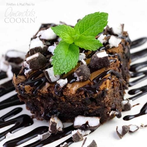 A close up photo of a peppermint patty brownie topped with drizzled chocolate sauce, chopped peppermint patties and a mint leaf.