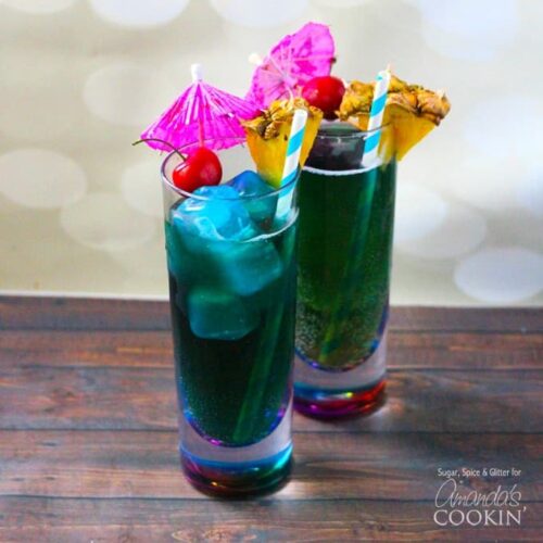 Two tall glasses filled with blue mermaid cocktails and topped with a cherry, pineapple wedge, an umbrella and a straw.